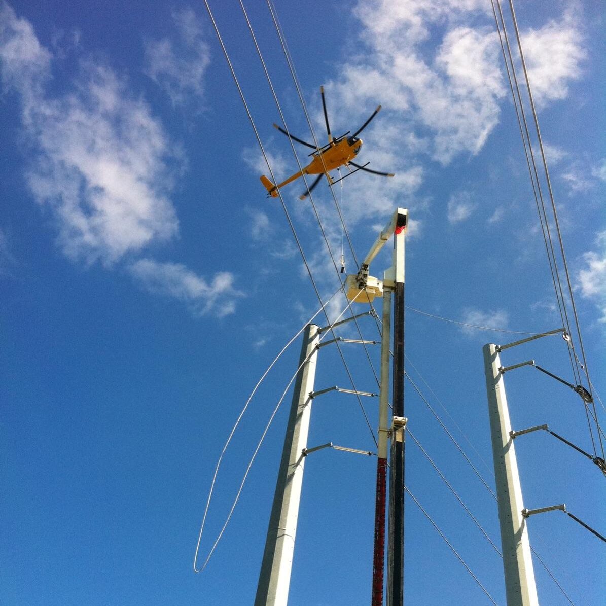 Yellow helicopter flying above tall electrical transmission towers and cables against a clear blue sky.